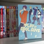 Ein Bild von einem Manga Regal. Im Vordergrund steht der erste Band von Play It Cool, Guys. Das Cover von diesem Manga ist in einem hellen blau gehalten, mit dem Schriftzug des Titels und vier Jungs auf dem Cover.
