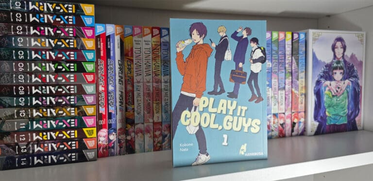 Ein Bild von einem Manga Regal. Im Vordergrund steht der erste Band von Play It Cool, Guys. Das Cover von diesem Manga ist in einem hellen blau gehalten, mit dem Schriftzug des Titels und vier Jungs auf dem Cover.